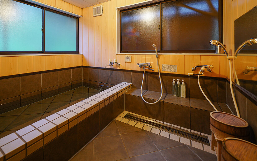 灰色のタイル貼りの浴槽、壁は木貼り、洗い場には木製の椅子と桶が置かれている